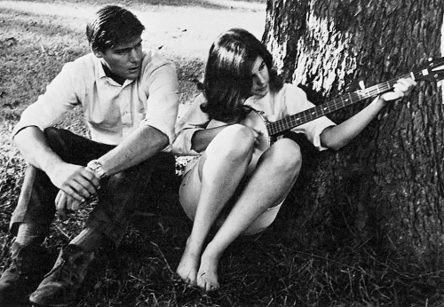 a woman plays a guitar underneath a tree while a man sits next to her, listening