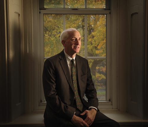 An older man sits in a window sill and is wearing a suit. 