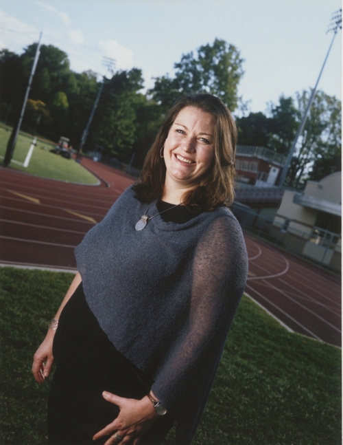 Kelly Wilcox is a woman standing next to a outdoor running track and smiling. 
