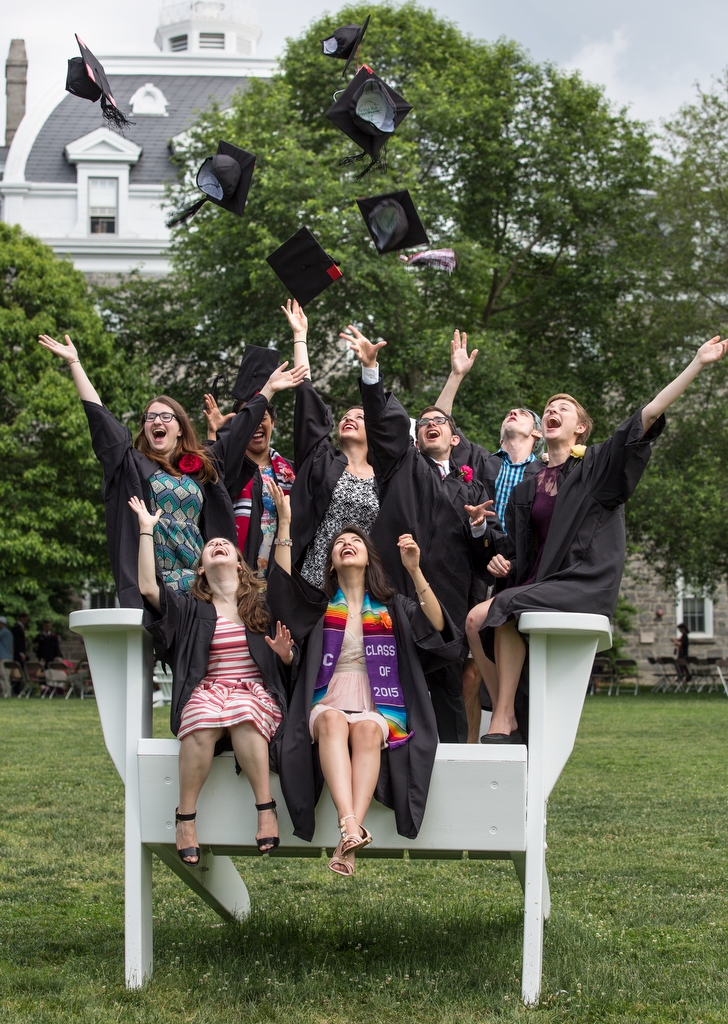 A group of seven swarthmore grads sit on the college's big adiorondak chair, their caps tossed, are suspended above them. 