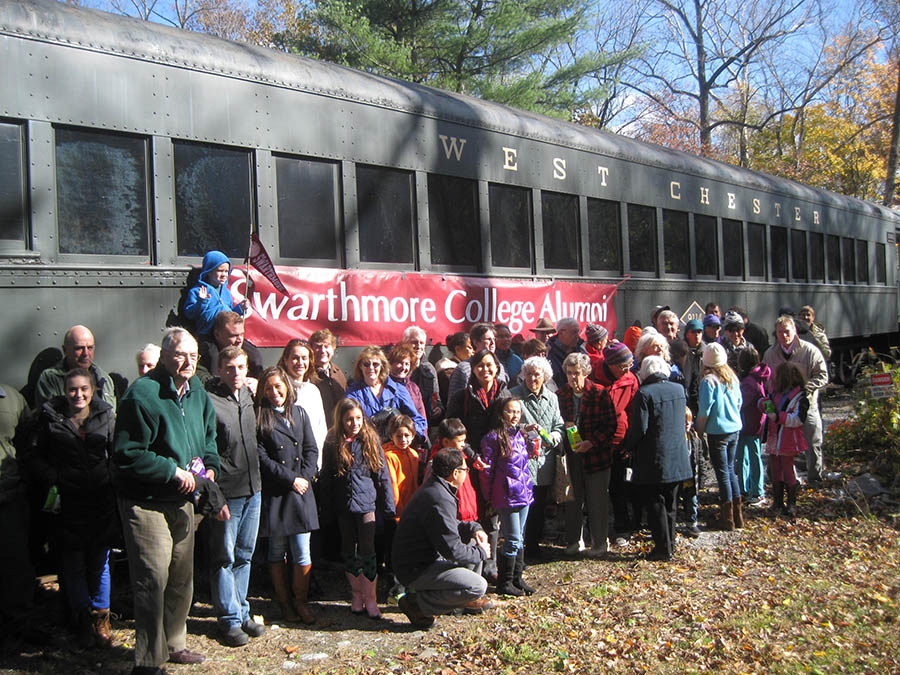 A group of people stand in front of a train outside with blue sky.