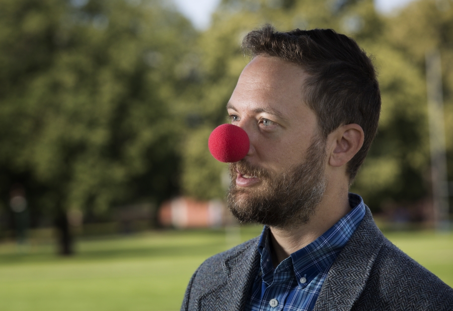 John Rieffel wears a clown nose and stands in a wooded area of campus