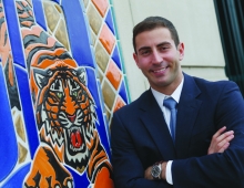 Sam Menzin ’12, in a suit, in front of the Detroit Tigers logo.