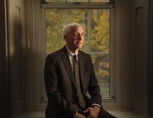 An older man sits in a window sill and is wearing a suit. 