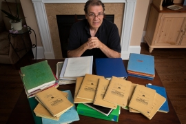 Matt Zencey ’79 sits in a well furnished home in front of a fireplace with his college notebooks scattered before him.