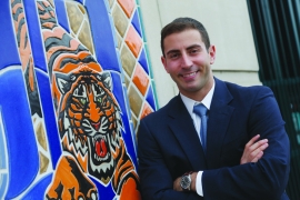 Sam Menzin ’12, in a suit, in front of the Detroit Tigers logo.