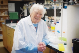 An elderly woman scientist is standing in a lab.