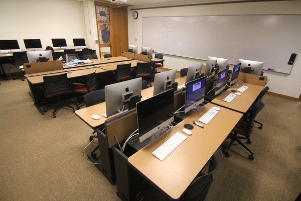 4 rows of desks and computers on retractible arms, 2 large whiteboards are fastened to the walls and a teaching podium is positioned in off to the side of the whiteboards.