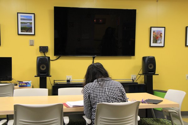 A large display screen is mounted on the wallabove 2 speakers. Student art is displayed on the walls. A person with long hair sits at a table studying.