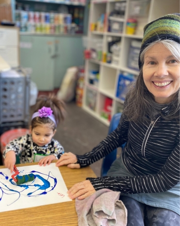 Smiling woman in the foreground sits at a table next to a child who is drawing.