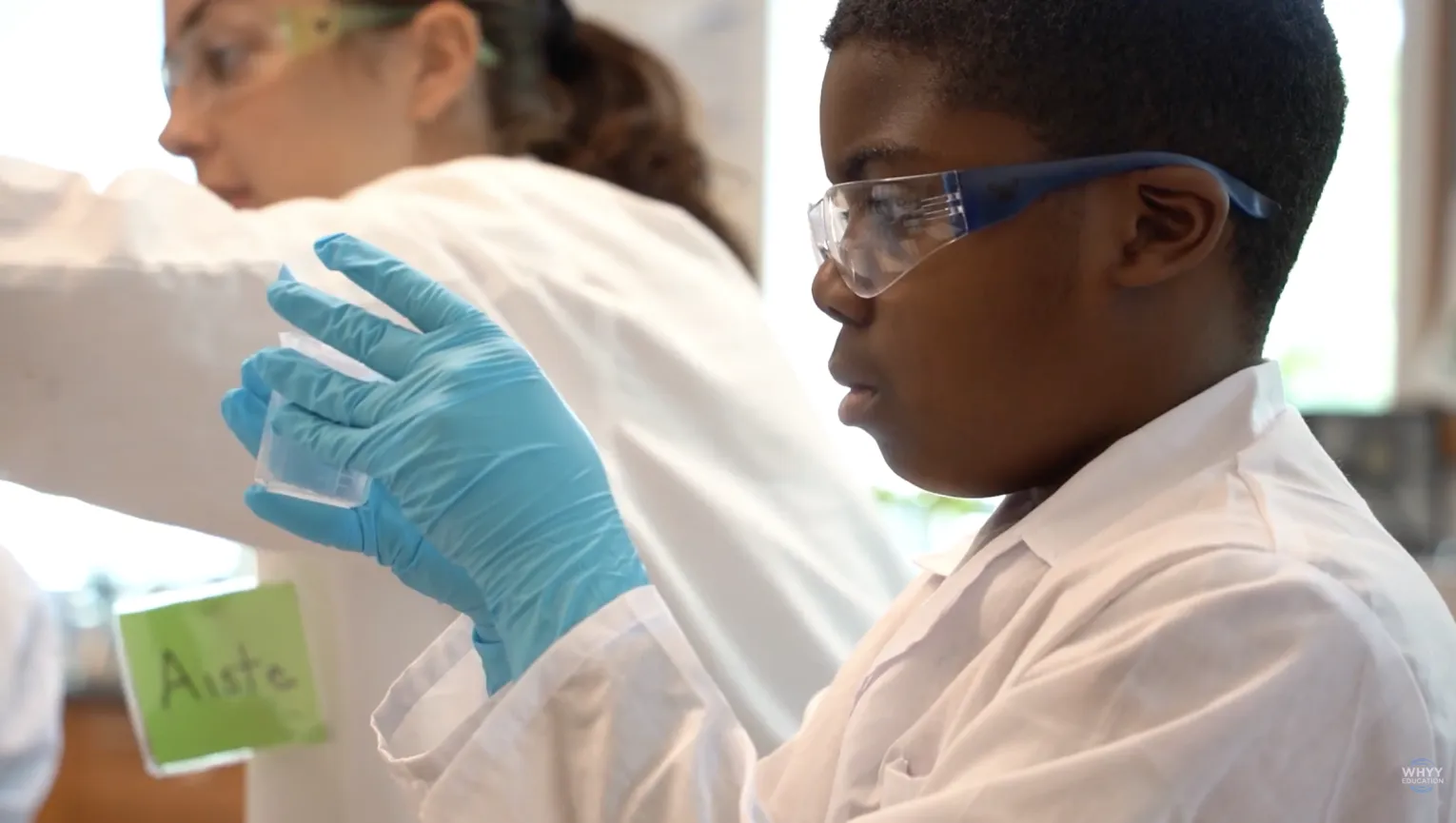 young boy in a white lab coat and plastic gloves holds a test tube with a college student teaching nearby