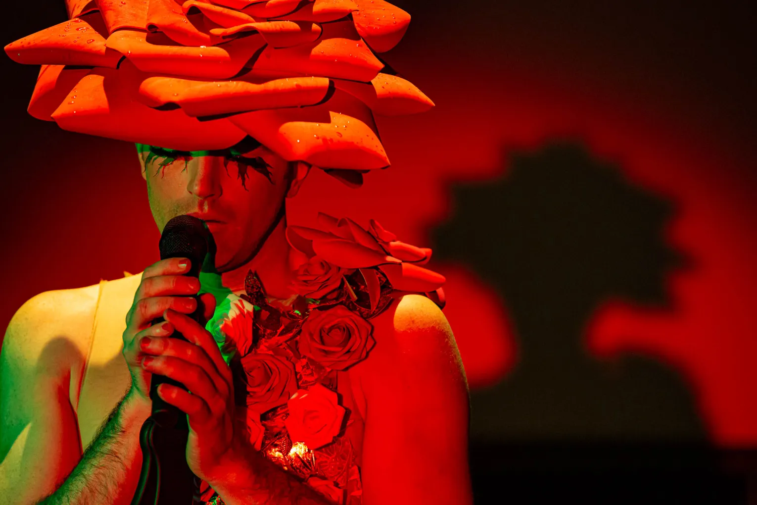 John Jarboe speaks into a microphone, wearing a headpiece that looks like a red rose.