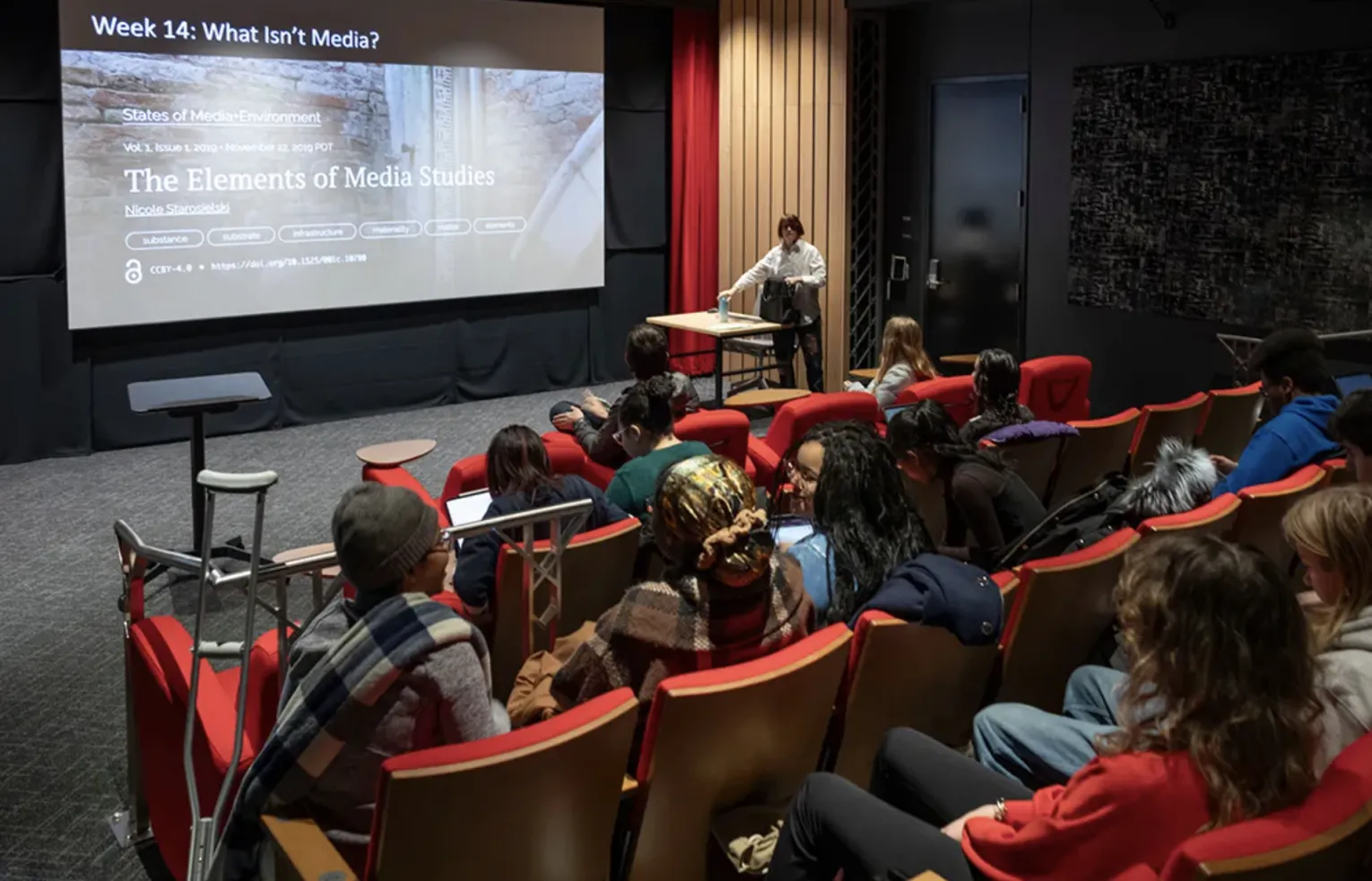 students sit in red cushioned chairs in Martin 028 Ping and Rose Chen Screening Room. Professor Patricia White is at the front of the room and has a slide up on the screening that reads Week 14 What Isn't Media