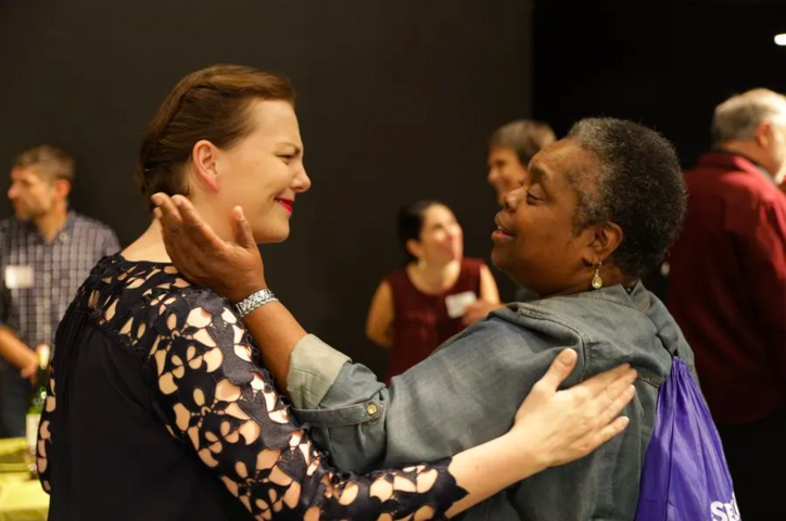 Two women embrace each other fondly in a rehearsal room. 