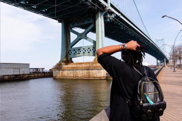 A person with dreadlocks and a backpack stands with their back to the camera and a hand on their head, looking up at the Benjamin Franklin Bridge.