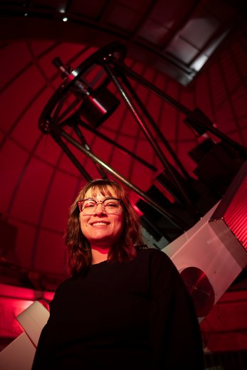 Student wearing glasses stands in front of large telescope in observatory