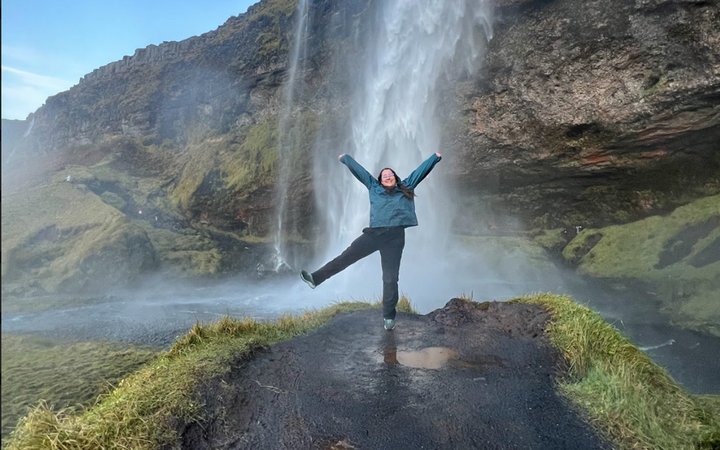 Person posing with arms and leg in the air in front of waterfall and mossy rocks