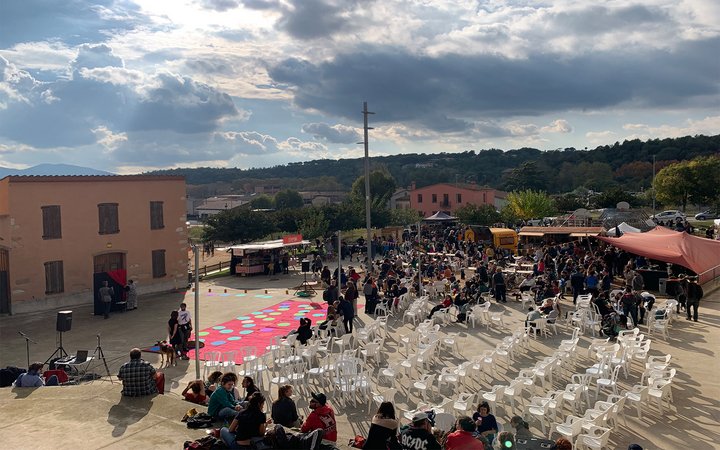 Crowd outside in large paved area with chairs and red mat for performers, with buildings and mountains in the background
