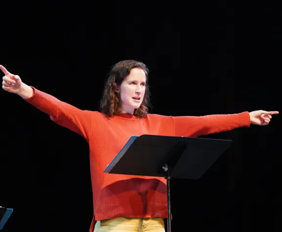 Rose Palmieri ('24) wearing a red shirt and pointing to the left and right in front of a music stand 