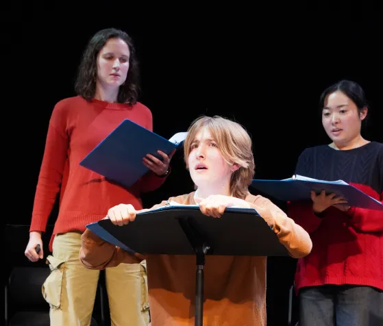 Rose Palmieri on the left standing behind Arlo Strauss who is kneeled before a music stand. Kana Nagata watches along from behind and to the right