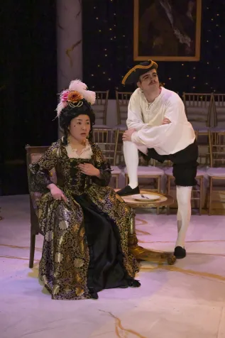 On the left, Kana Nagata is dressed in an 18th Century Gown with her hand to her heart. To her right is Griffin Moore, wearing a tricorn hat, white shirt, and knickers. He has one foot up on a table 