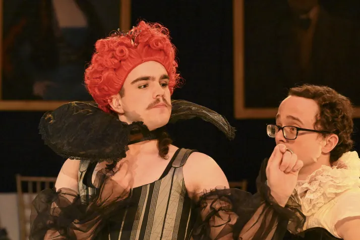 Griffin Moore is wearing a pink wig and a corset. He makes a dismissing gesture. To his left is Frank Moore, wearing a lace collar. 