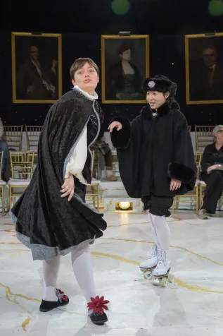 Lizzie Culp looks at the camera with a quizzical expression, and flipping a cape behind her. Kana Nagata looks on from the side, wearing roller skates.