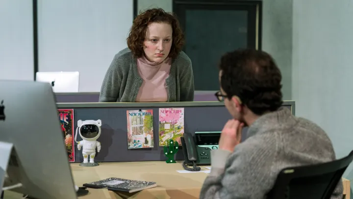 Gloria, a woman in a grey cardigan, glares at Sasha, a person sitting behind a desk 