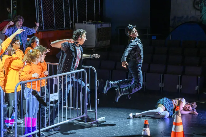 Macheath stands behind bike racks with his knife pulled back while Captain Smith jumps after just having been stabbed in the rear end