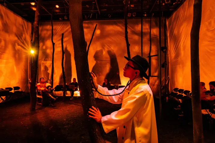 A still from Swarthmore's Woyzeck. A doctor in a dark hat holds a stehoscope up against a tree