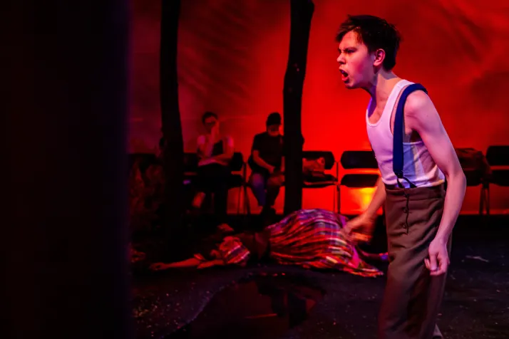 Woyzeck screams against red light. He is wearing a white tanktop with suspenders and brown pants 