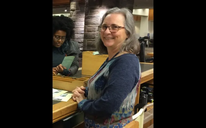 A woman with shoulder-length grey hair and glasses wearing a dark top smiles over her shoulder at the camera.