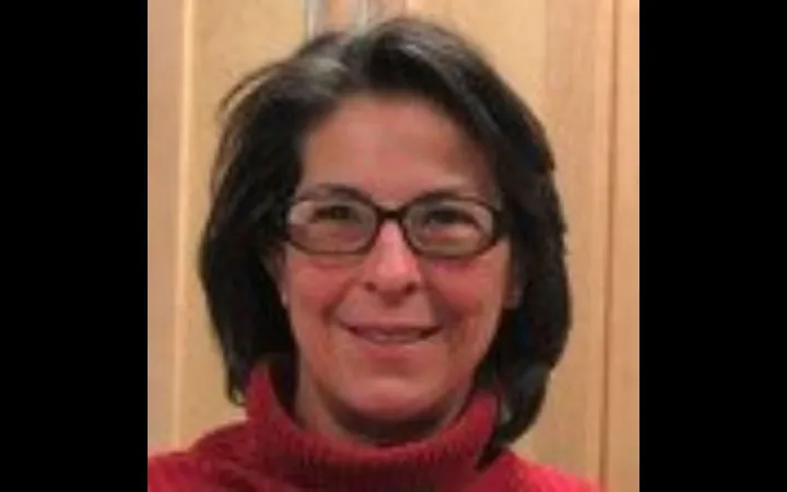 Close shot of a woman with shoulder length grey hair wearing glasses and a red turtleneck, smiling at the camera.