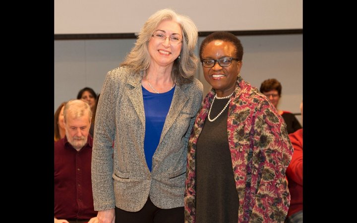 Two woman stand beside each other, smiling at the camera. The woman on the left wears glasses, a grey jacket, and a bright blue top and has shoulder length white hair. The woman on the right has close cropped hair and wears glasses and a long patterned jacket.