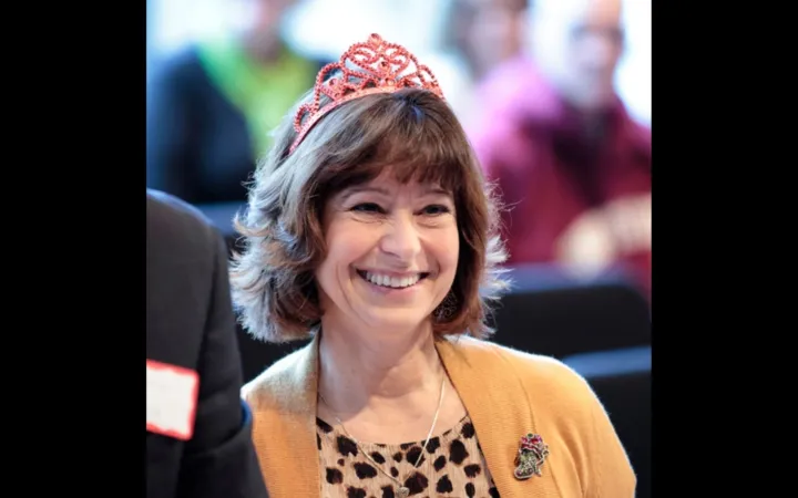 A woman with brown hair and bangs wearing a yellow cardigan and pink toy tiara smiles widely.