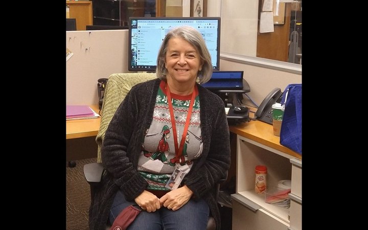 Woman with short grey hair wearing a long black cardigan and red lanyard sits at a desk with a computer behind her and smiles at the camera.