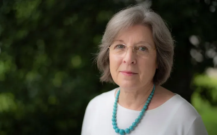 Outdoor headshot of a woman with shoulder length grey hair and glasses wearing a white top and turquoise necklace.