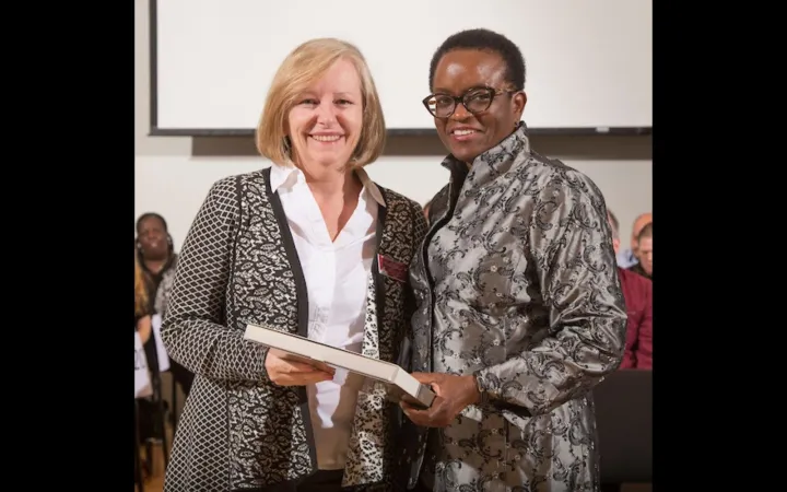 A woman with blonde hair and bangs wearing a white button down shirt and black patterned cardigan stands next to a woman with close cropped dark hair and glasses, wearing a patterned grey jacket. Both smile at the camera.