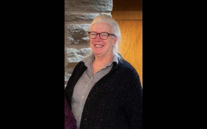 A woman with white hair and thick dark glasses, wearing a grey shirt and black cardigan, smiles at the camera.
