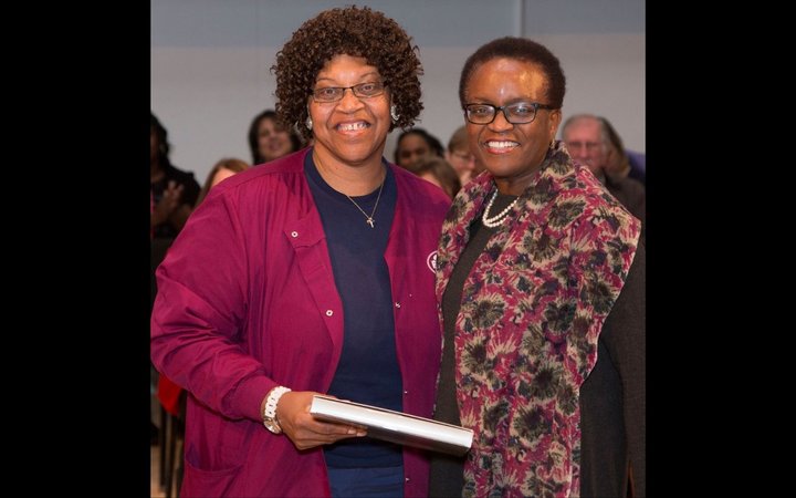 Two woman stand beside each other and embrace, smiling at the camera. The woman on the left wears a garnet jacket and navy top and has shoulder length hair. The woman on the right has close cropped hair and wears glasses and a long patterned jacket.