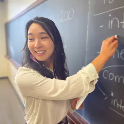 Asian American woman with long black hair smiles and looks off camera while writing on the chalkboard in a classroom