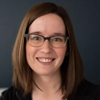 Caroline Larsen, a young white woman with dark hair and glasses, smiles in front of a dark background.