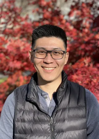 An Asian male wearing glasses, smiling in front of a Japanese maple tree.