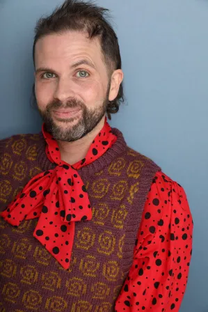 Connor Hogan's headshot. A nonbinary person wearing a polka-dot pussy bow tie blouse and a purple vest with yellow spirals
