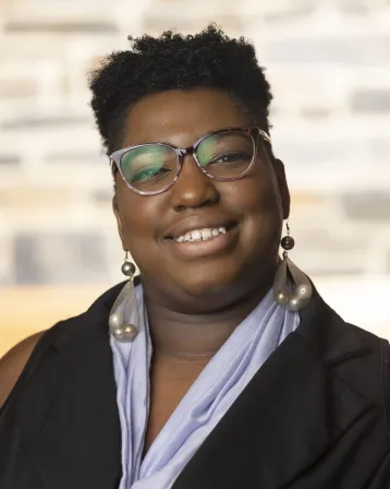 A black femme-presenting person with a black and lavender top, wearing glasses and large dangle earrings