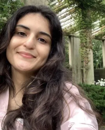 A young woman with long dark hair smiles at the camera in a close-up outdoor portrait. She is wearing a light pink top, with leafy greenery and a concrete architectural structure in the background.