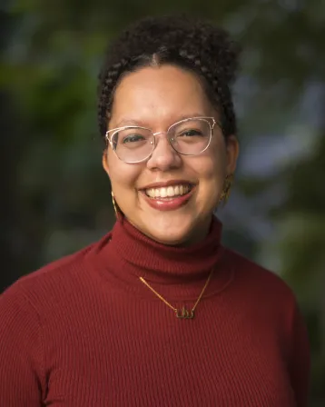 A light-skinned Black woman wearing gold glasses and a red turtleneck sweater smiles at the camera.
