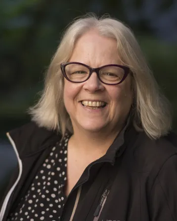 Sunka Simon headshot in which she wears a black shirt with white polk a dots with a black jacket over top and smiles. Greenery is visible in the background