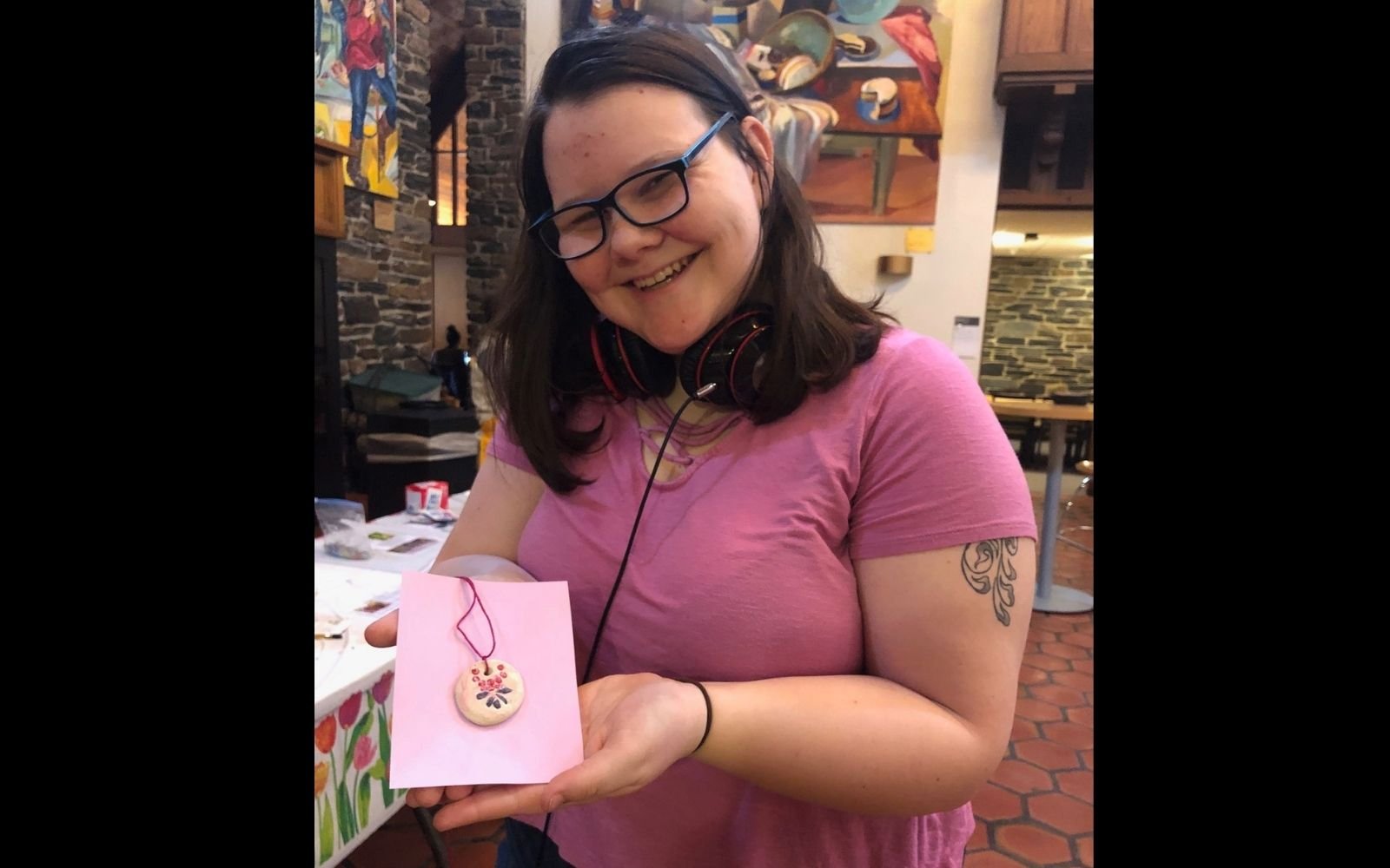 A smiling person with brown hair, glasses, and a pink tee shirt holds up a ceramic necklace to the camera