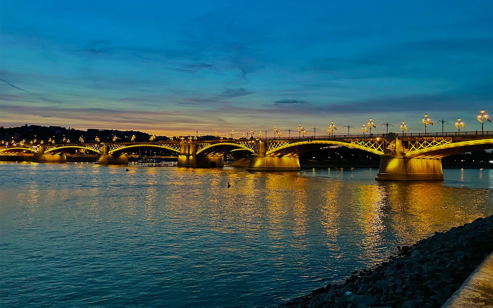 Bridge over large body of water at dusk, with bright yellow lights that are visible in reflection